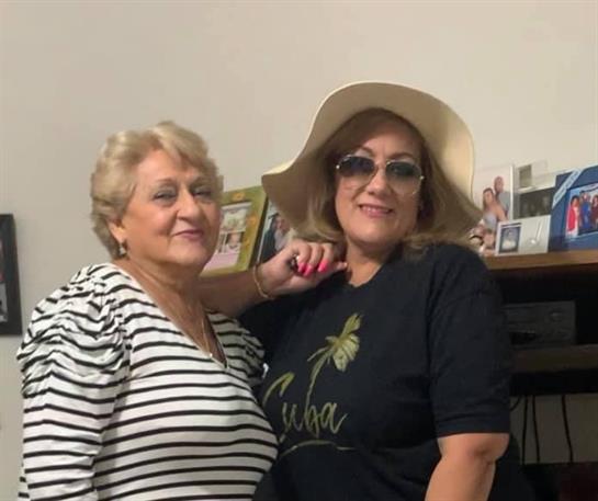 Two women with sunglasses and hats smile while posing warmly in a cozy living space.
