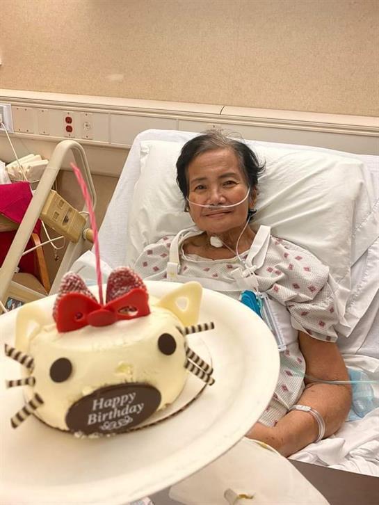 An elderly woman in a hospital bed enjoys a birthday cake with a cat design and smile.