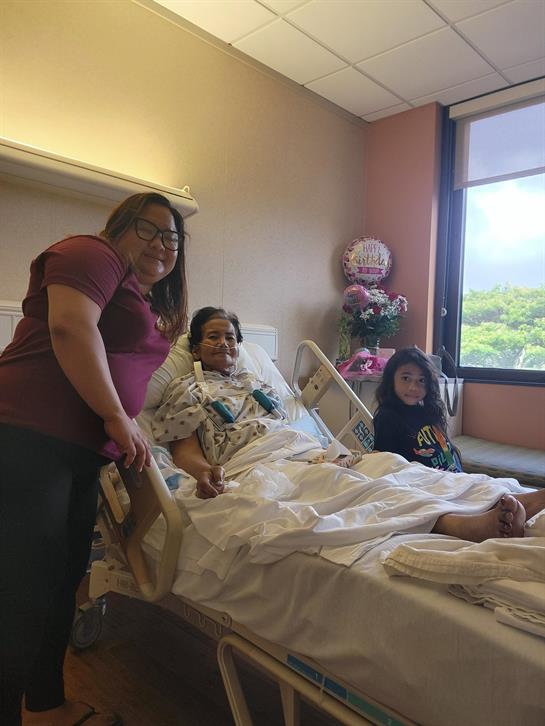 A woman is visiting a patient in a hospital room with a child nearby, providing support and comfort.