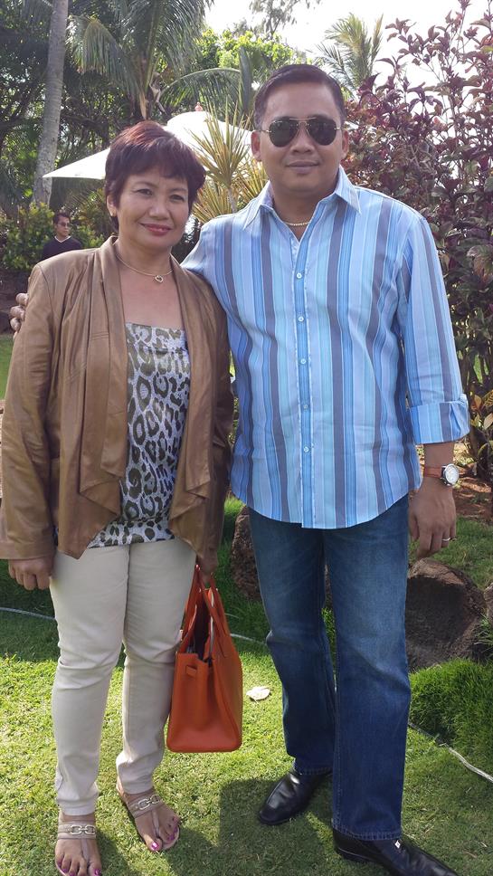 Two people stand together in a beautiful garden, smiling and dressed in casual attire.