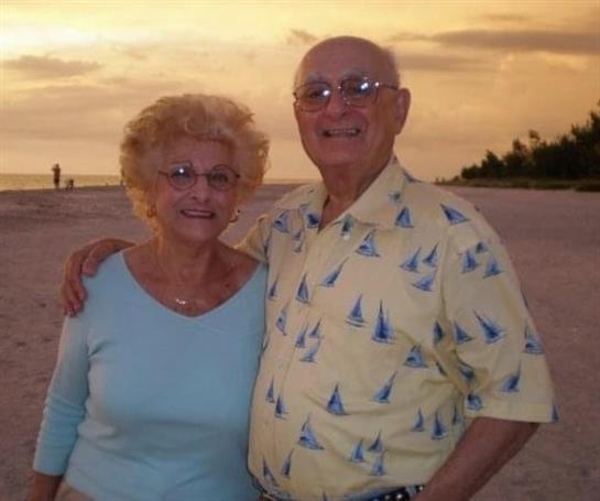 A joyful elderly couple smiles together on a beach during a vibrant sunset.