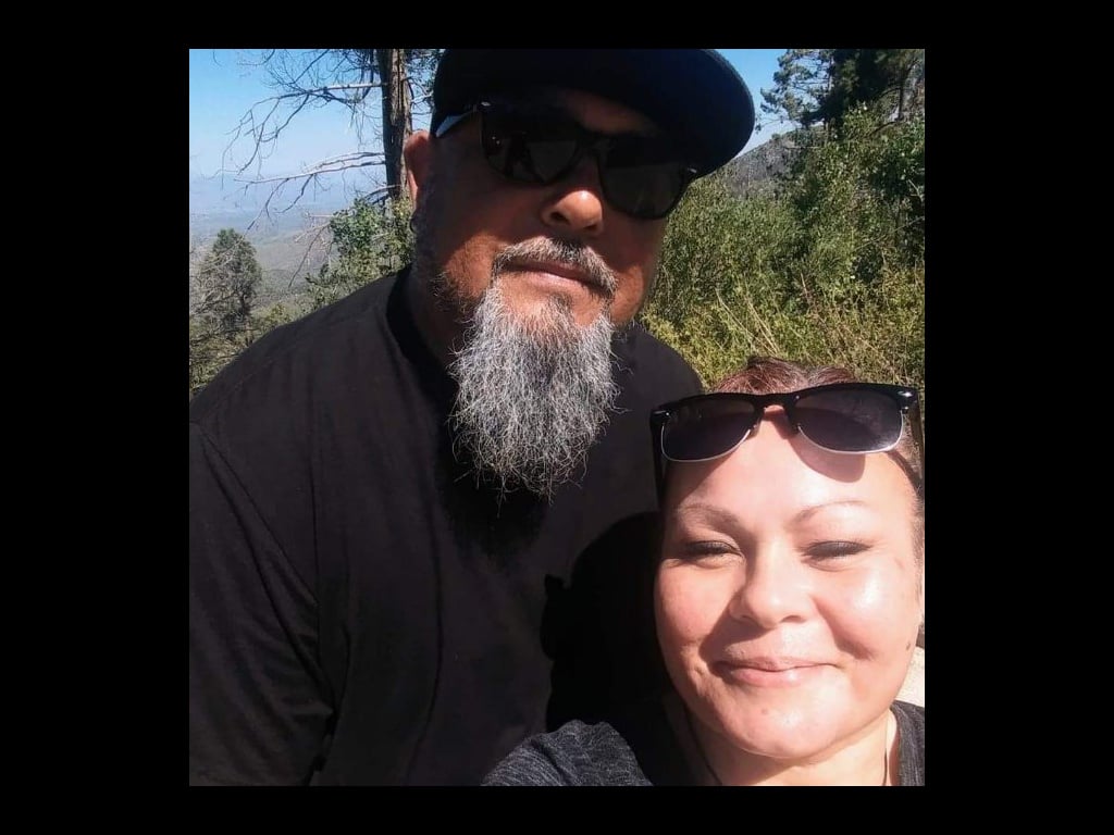 A man and woman take a selfie together while hiking in the mountains under clear skies.
