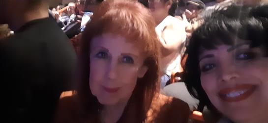 Two women pose for a selfie while attending a vibrant event in a packed venue.