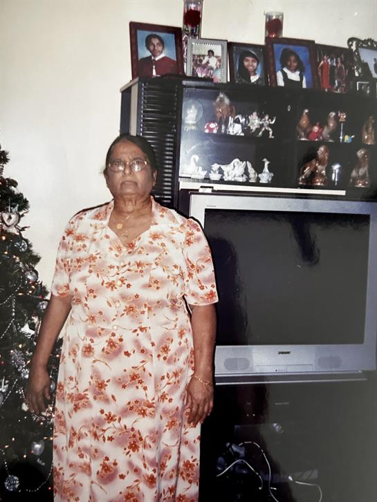 A woman in a floral dress stands in her living room next to a decorated Christmas tree.