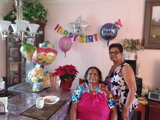 Family celebrates a birthday in a decorated room with balloons and plants present.