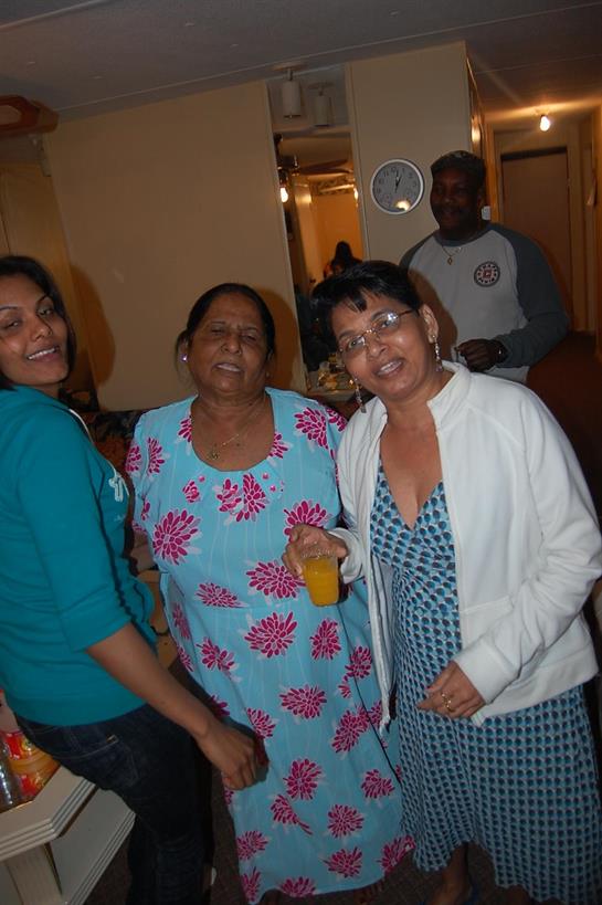 Three women share smiles and laughter, with one holding a drink, in a cheerful atmosphere.
