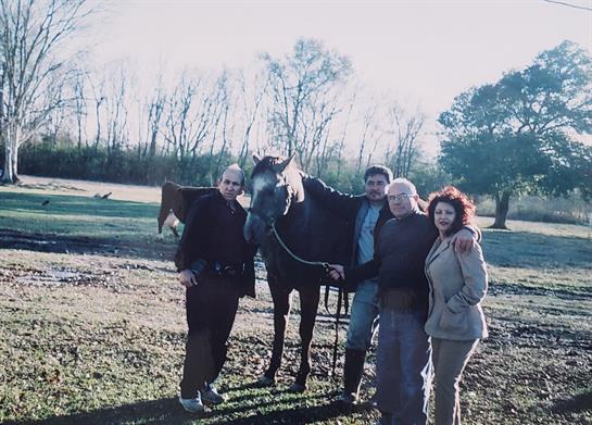Friends gather around a horse in a sunny, open area surrounded by trees.