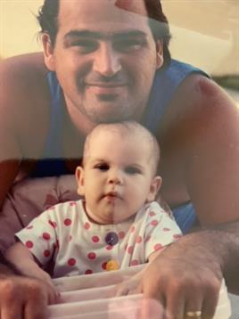 A father smiles at the camera while holding his baby, both enjoying a delightful moment outdoors.