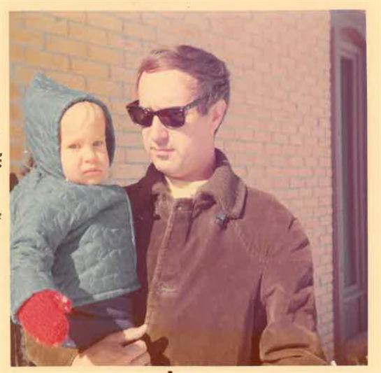 A man wearing sunglasses holds a child dressed warmly in a quilted jacket with mittens.