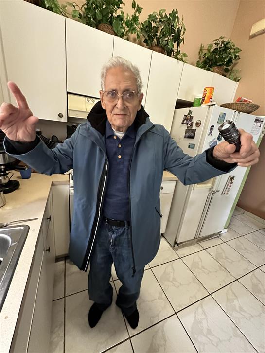 An elderly man stands in a kitchen, gesturing playfully with both hands while wearing a coat.