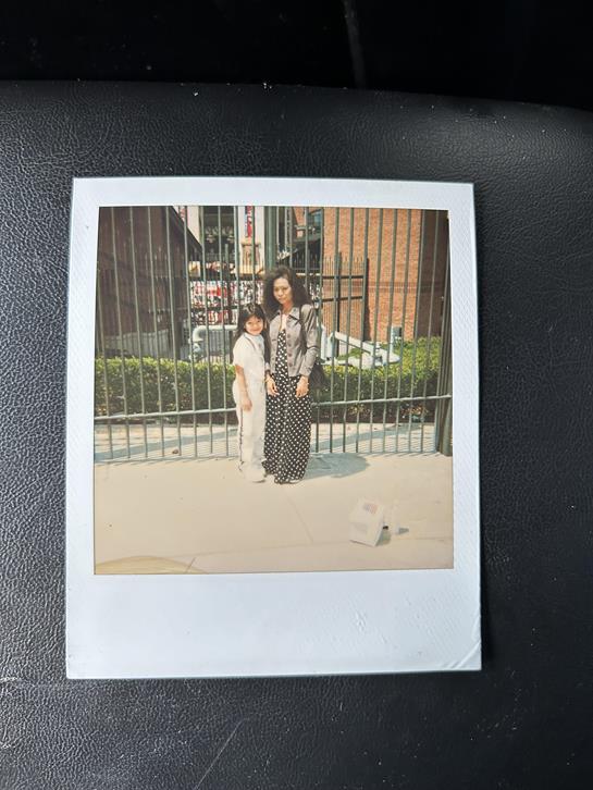 A woman and a young girl stand side by side outside near a fence, smiling.