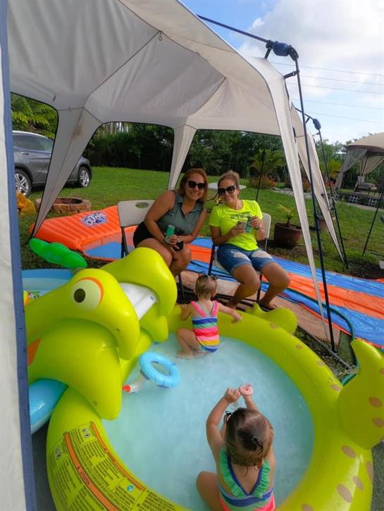 Two adults relax under a tent while children play in a colorful pool on a sunny day.