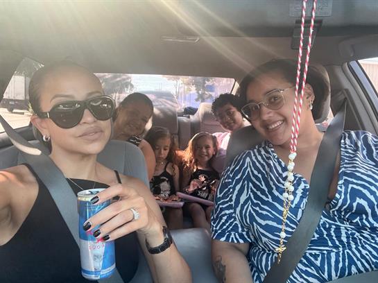 A group of adults and children share smiles and drinks during a car ride in daylight.