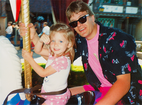 A young girl smiles widely while riding a carousel with her father in vibrant attire.
