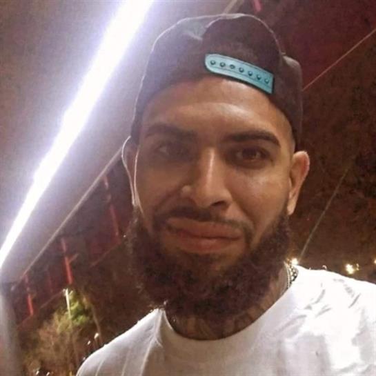 A man with a beard and cap stands under urban lights at night, smiling casually.