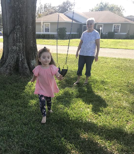 A cheerful girl runs toward a swing as her grandmother looks on with a smile in the park.