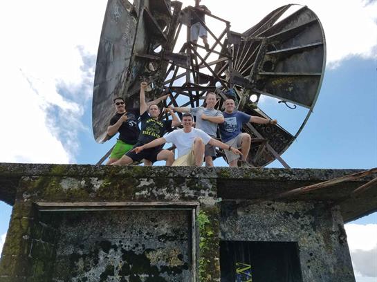 Five friends are joyfully posing on a rooftop next to a large satellite dish on a sunny day.