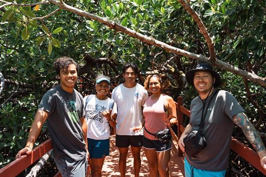 Friends smile and pose together on a bridge surrounded by lush greenery in bright sunlight.