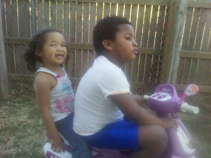 Two kids share a toy motorcycle, laughing and having fun in the backyard.