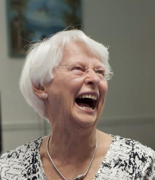 An elderly woman with short white hair laughs joyfully at a gathering.