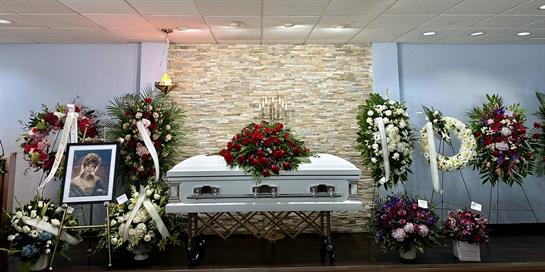 A memorial service features a casket surrounded by vibrant floral arrangements and a portrait.