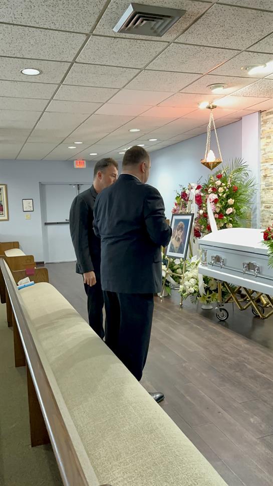 Two men stand solemnly beside a casket, honoring the memory of a loved one in a quiet chapel.