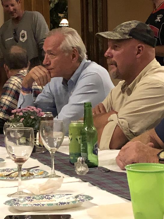 Two men engaged in conversation at a dinner table surrounded by friends and drinks.
