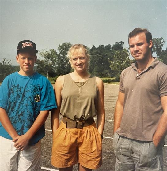 Three individuals gather in a park on a warm day, smiling while standing close to each other.