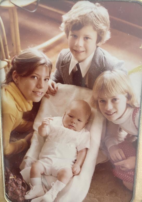 Four children share smiles and warmth, with one cradling a baby in a bright indoor space.