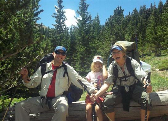 Family members relax on a log while hiking in a wooded area under bright sunshine.