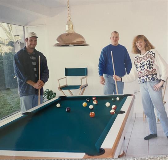 Three friends gather around a pool table, smiling while holding cues in a bright room.