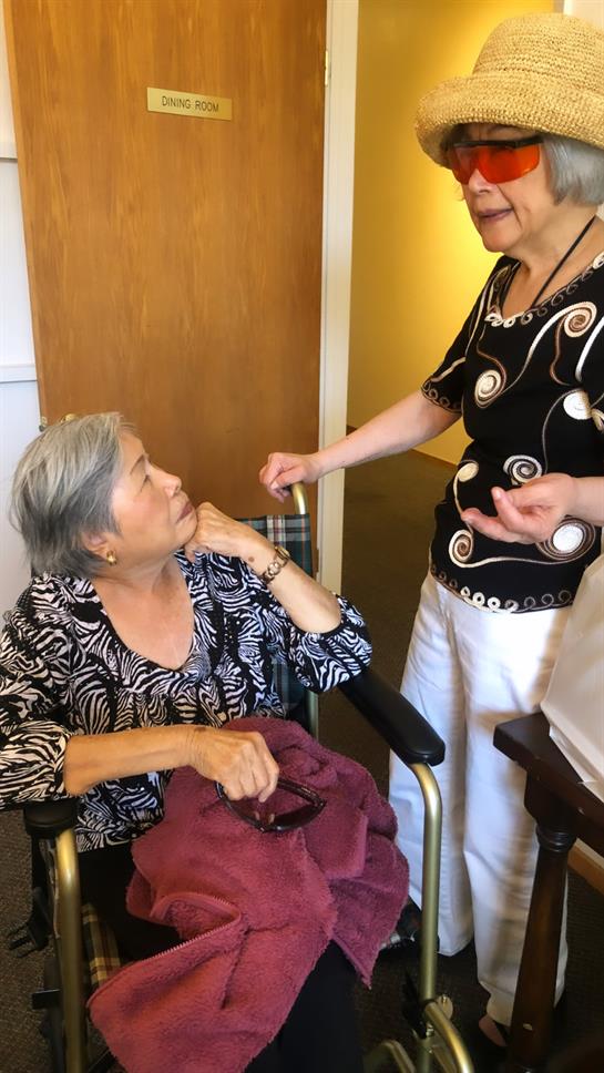 Two women in a care facility share lively conversation while seated and standing.