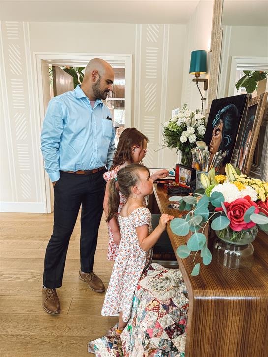 A man and two children engage with a colorful artwork in a bright, floral room.