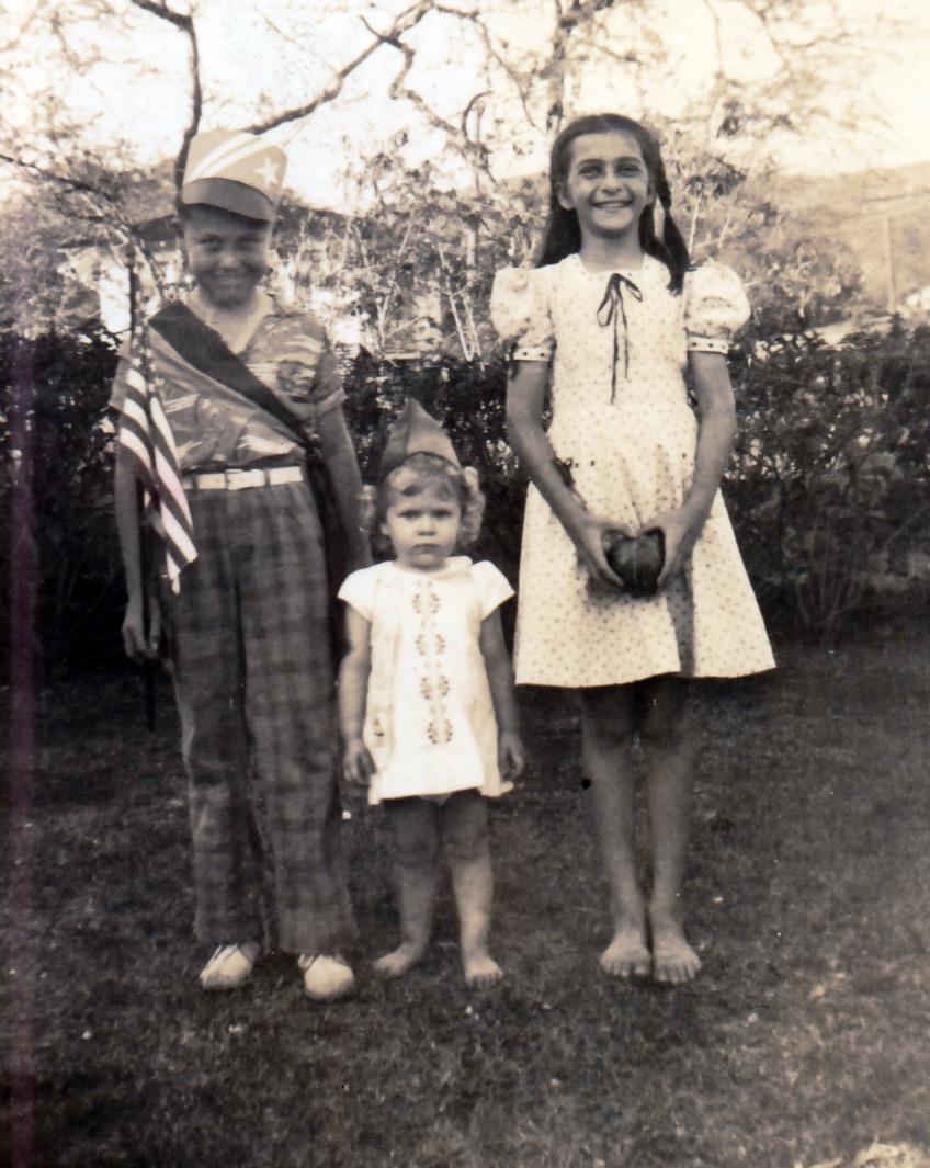 Three children stand together in a sunny garden, adorned in festive outfits and symbols.