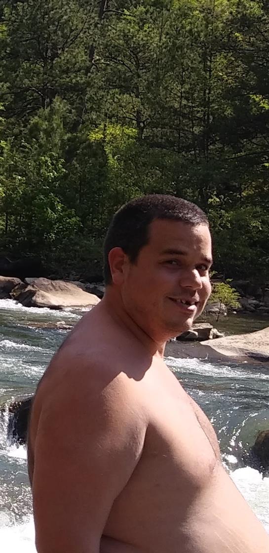 A man stands by a clear river, smiling. Sunlight shines through green trees in the background.