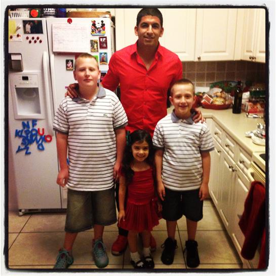 Children wearing matching shirts stand with an adult in a lively kitchen setting.