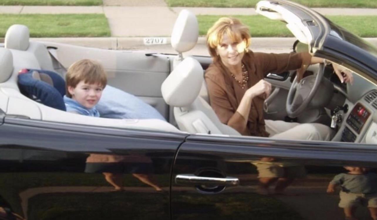 A woman drives a convertible with a child next to her, both smiling and enjoying a sunny day.
