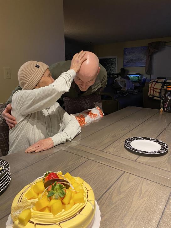 Two friends smile and embrace while enjoying a casual gathering with cake on the table.