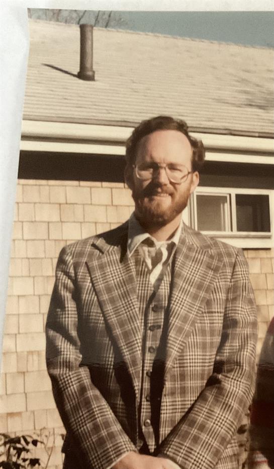 A man wearing a plaid suit and glasses stands confidently outside a vintage building.
