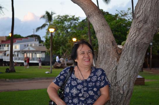 A woman smiles while standing next to a large tree in a park during the early evening.