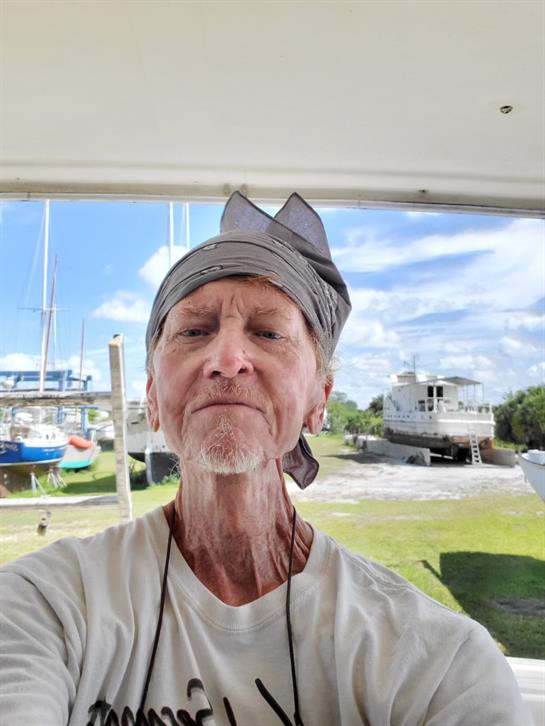 Man with a bandana smiles for a selfie at a marina with boats and blue sky.