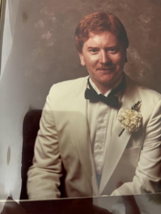 A groom wears a white tuxedo and black bow tie, smiling warmly at a wedding celebration.