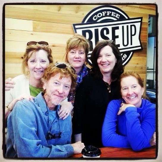 Five friends gather around a table, smiling and sharing laughter in a warm coffee shop setting.
