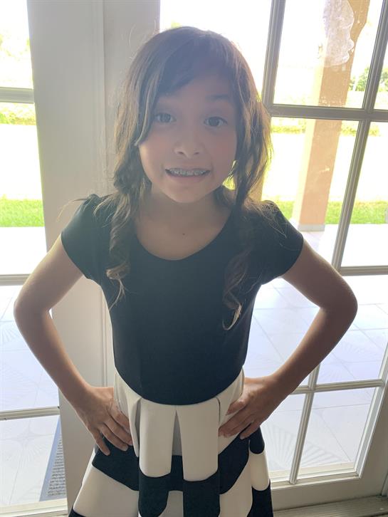Girl with long hair stands with hands on hips, smiling happily by a sunny window.