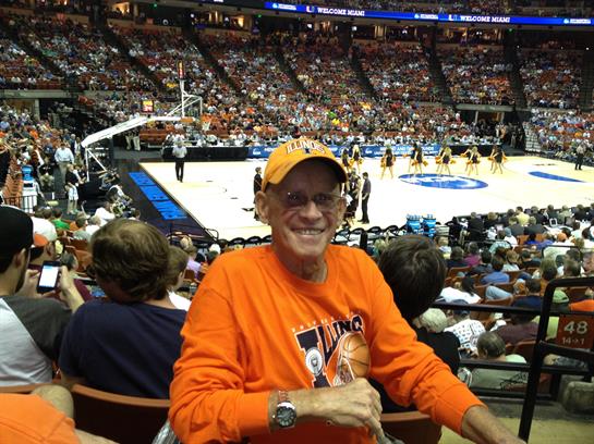 A delighted fan in orange attire smiles while sitting among other spectators at a basketball game.