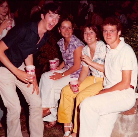 Friends gather together at a lively outdoor party, holding drinks and smiling joyfully.