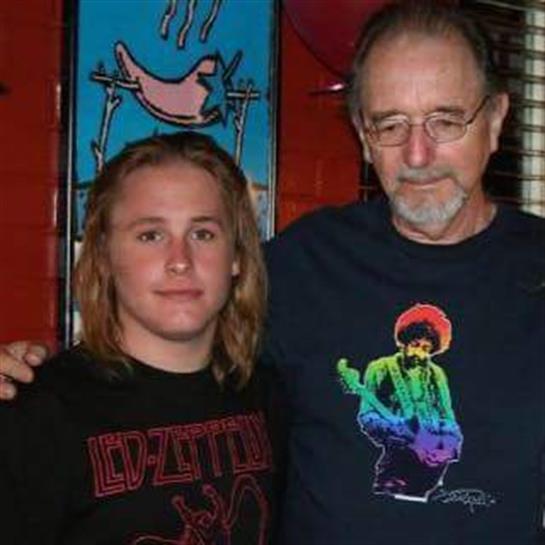 A young man with long hair and an older man with glasses smile in a lively setting.