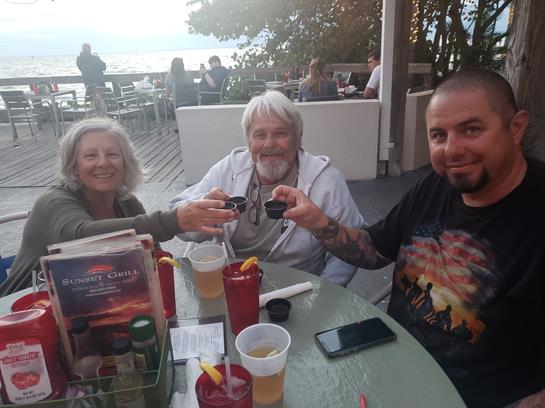 Three friends raise their drinks in celebration at a seaside terrace during sunset.