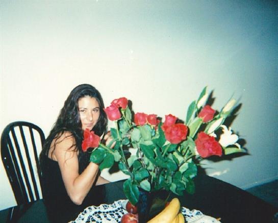 A woman sits at a table with red roses and bananas, appearing relaxed in a cozy indoor space.