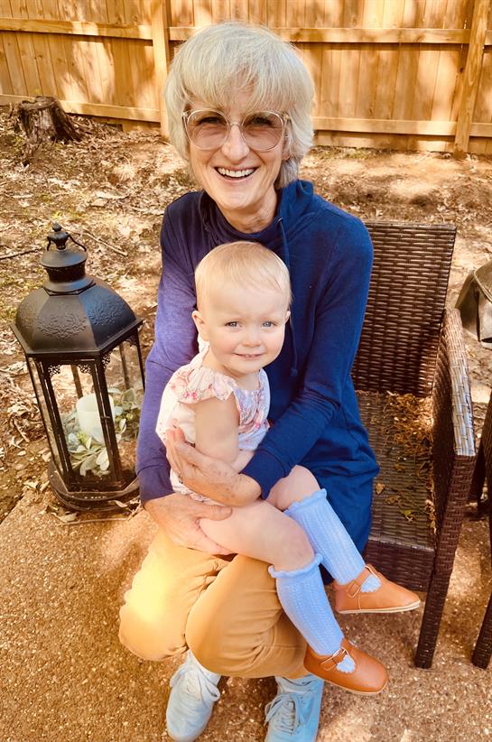 A grandmother smiles brightly while holding her granddaughter on her lap outside in nice weather.